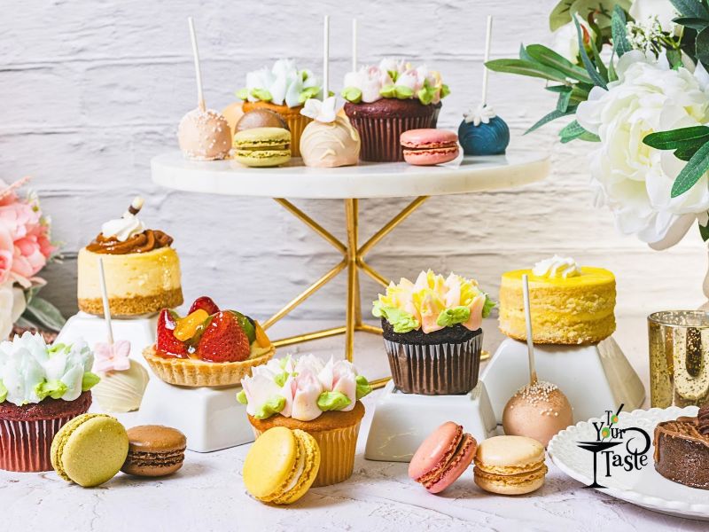 Photo of a dessert table. Cake pops, cookies, mini cheesecakes, and fruit tarts. There are some flowers to the left and right - the flowers on the left are pink and on the right are white. The color palette is in pastels. The table and background are white.