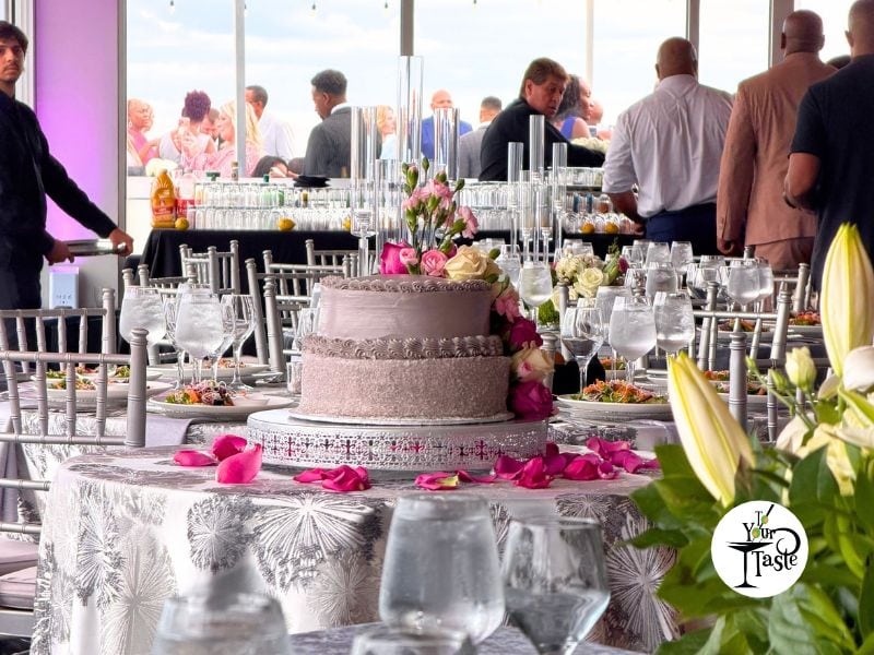A photo at an event with white tablecloths with pale dark flower outlines. There is a wedding cake with pale brown frosting and pink flowers on top and around it in the center of the image. There are glasses and flowers and people walking around and a wall of glass that looks out to the sky.